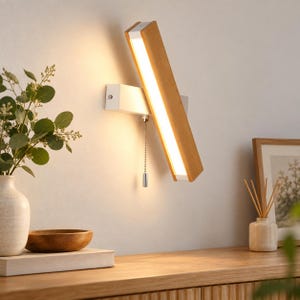 May include: A wall-mounted light fixture featuring a rectangular wooden frame with an integrated LED light strip. The fixture is attached to a white base and includes a silver pull chain switch hanging vertically from the center of the unit.
