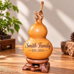 May include: A tan, bulbous, dried gourd with a natural stem top, resting on a carved wooden base. The gourd features dark brown engraved text that reads Smith Family and EST. 2026, centred on the rounded body of the smooth, organic-shaped object.
