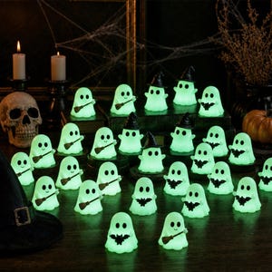 Op de afbeelding: Een verzameling kleine, lichtgevende lichtgroene spookfiguurtjes op een donker oppervlak. Elk spookje heeft een eenvoudig lachend gezicht en houdt een miniatuur zwarte heksenhoed, een kleine bruine bezem of een kleine zwarte vleermuis vast.