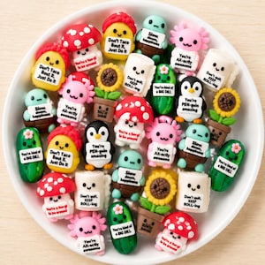 May include: A collection of small, colourful clay figurines in a white bowl. The figures include red mushrooms, green pickles, pink axolotls, penguins, sunflowers in pots, and toilet paper rolls, each holding a sign with punny text like Don't Taco Bout It.