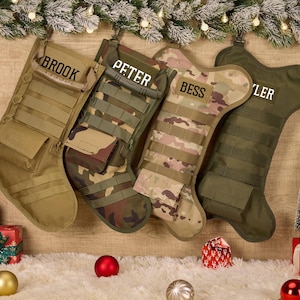 Puede incluir: Cuatro calcetines navideños tácticos colgados en fila. El primero es de color tostado con el nombre BROOK, el segundo es de camuflaje boscoso con PETER, el tercero es de camuflaje multiterreno con BESS y el cuarto es verde oliva con TYLER.