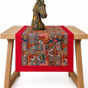 Op de afbeelding: Een rechthoekige tafelloper met een centraal paneel met een veelkleurig geometrisch en bloemmotief in tinten rood, blauw, geel en wit, omrand door een effen rode stoffen rand, met daarop een donkerbruin sculptuur van een paardenkop.