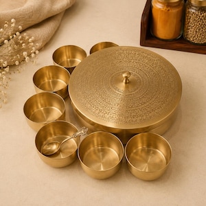 Könnte beinhalten: Eine runde Gewürzdose aus Messing mit einem aufwendig gravierten Deckel und einem zentralen Knauf, umgeben von sieben kleinen passenden Messingschalen. Eine Schale enthält einen kleinen Messinglöffel. Das Set hat ein poliertes Metallfinish.
