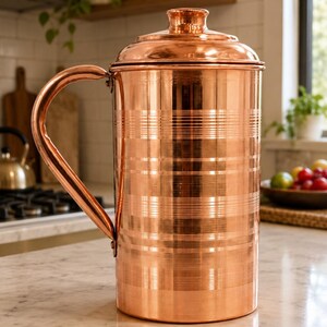 May include: A polished copper pitcher with a cylindrical body and a matching lid. The exterior features horizontal bands of fine etched lines encircling the vessel. It includes a curved handle attached to the side, showcasing a metallic finish.