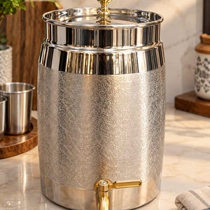Op de afbeelding: Een cilindrische roestvrijstalen drankdispenser met een gepolijste afwerking, een gestructureerd damastpatroon op de behuizing, een goudkleurige dekselknop en een bijpassende goudkleurige kraan aan de onderkant.
