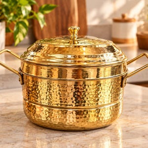Può includere: Una pentola a vapore in metallo martellato color oro con due manici laterali e un coperchio abbinato dotato di pomello centrale. La pentola presenta un design a gradini con una superficie testurizzata e incavata che riflette la luce.