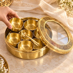 Puede incluir: Un recipiente redondo de metal dorado con una tapa de cristal transparente que presenta un borde grabado ornamentado. Dentro del recipiente hay siete cuencos pequeños de metal dorado a juego y una pequeña cuchara de metal dorado encima.