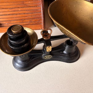 Op de afbeelding: Een zwarte gietijzeren balansweegschaal met twee koperen schalen. De linker schaal bevat een stapel zwarte gewichten, waarbij het bovenste gewicht is gemarkeerd met 1 OZ. Een centraal koperen mechanisme verbindt de schalen. Een zwart label toont SALTER No 50 TRADE MARK.