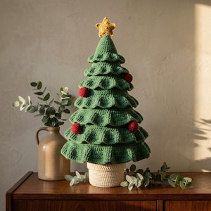 May include: A crocheted Christmas tree in shades of green with ruffled tiers, topped with a yellow star. The tree is decorated with small red crocheted spheres and sits in a beige crocheted pot. The texture is visible throughout the yarn work.