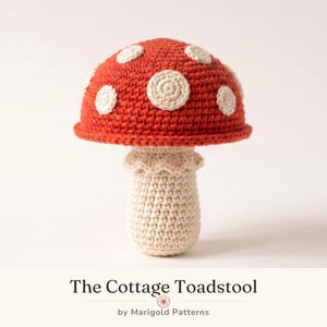 May include: A crocheted mushroom toy featuring a vibrant red cap adorned with circular cream-colored spots and a textured cream-colored stem with a ruffled collar. The text below reads The Cottage Toadstool by Marigold Patterns.