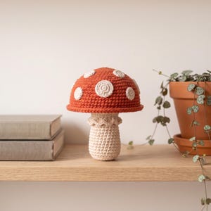May include: A handcrafted crochet mushroom featuring a vibrant orange cap adorned with circular white spots and a textured cream-colored stem. The base of the cap includes a delicate ruffled edge detail, creating a distinct, soft-textured appearance.