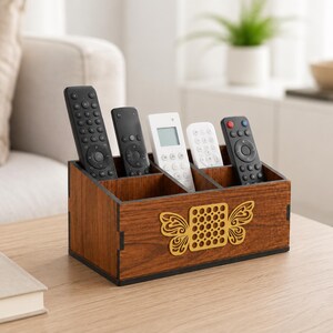 May include: A rectangular wooden remote control holder with four compartments. The front panel features a decorative gold-colored cutout design resembling a butterfly with a grid-patterned center. Five various remote controls are stored inside.
