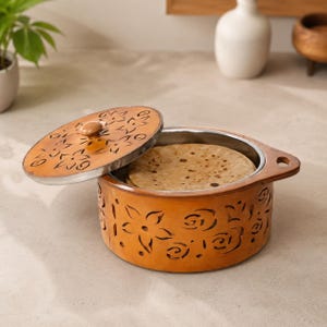 May include: A round wooden food container with a removable lid, featuring intricate floral cutouts on the sides and top. The interior is lined with stainless steel and contains several flat, round, light brown bread items inside the vessel.