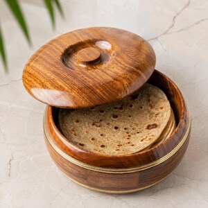 May include: A round wooden container with a matching lid, featuring a polished brown wood grain finish and thin brass inlay bands. The container holds a stack of round, flat, light brown unleavened bread discs with visible dark brown spots.