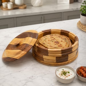May include: A round wooden container with a removable lid, featuring a checkerboard pattern of light and dark wood tones. Inside the container are several flat, round, golden-brown breads. Two small bowls containing white sauce and red food sit nearby.