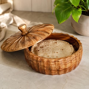 May include: A round wooden container with a carved, fluted exterior and a matching lid, containing a stack of flat, round, beige tortillas. The wood features a natural grain pattern with light and dark brown tones throughout the structure.