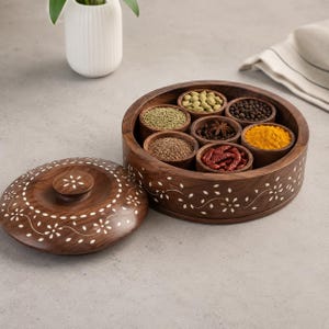 May include: A round wooden spice box with a matching lid, featuring white floral inlay patterns. The container holds seven small, removable wooden bowls filled with various colorful spices, including turmeric, dried chili peppers, and whole peppercorns.