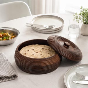May include: A round, dark brown wooden container with a textured, ribbed exterior and a matching circular lid, containing a flat, round, beige bread. The container features a small, centered knob on the lid for easy handling and removal.
