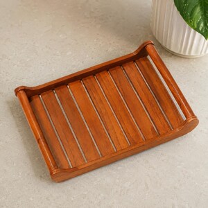 May include: A rectangular wooden tray featuring a slatted base design with evenly spaced brown planks. The tray includes two curved, elevated side handles that extend upward from the frame, providing a structured and functional appearance.