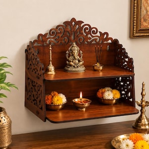 Peut inclure: Une étagère murale en bois brun foncé à deux niveaux avec des motifs découpés au laser sur les panneaux arrière et latéraux. L'étagère contient une statue de Ganesh en laiton, une cloche en laiton, de l'encens et deux bols de fleurs colorées.