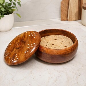 Puede incluir: Un recipiente redondo de madera de color marrón oscuro con tapa a juego, que presenta delicados patrones florales incrustados en blanco. El recipiente está abierto, revelando un pan plano circular de color marrón claro con veta.