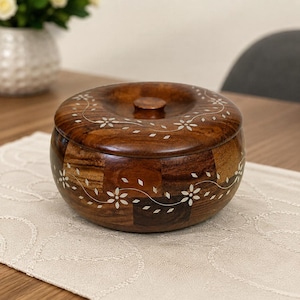 Puede incluir: Una caja redonda de madera marrón oscuro con tapa, que presenta un diseño de parches con vetas de madera e incrustaciones florales blancas delicadas que envuelven los lados y la superficie superior, con un pequeño pomo central circular.