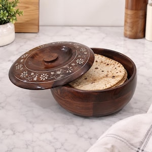 May include: A dark brown wooden bowl with a matching lid, featuring an inlaid floral pattern in a light-colored material. The bowl contains several round, flat, toasted bread items. The lid is partially removed, resting at an angle on the bowl.