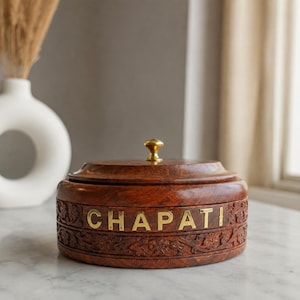 May include: A round, dark brown wooden container with a lid and a small gold-colored knob. The side features intricate floral carvings and the word CHAPATI in gold-colored metal lettering. The container has a smooth, polished finish throughout.