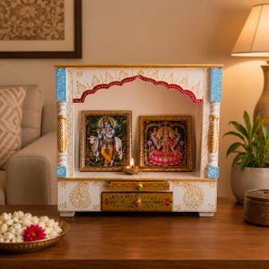 May include: A white wooden home temple featuring gold and blue painted accents, an arched top with red trim, and two drawers. Inside, two framed religious prints are displayed alongside a small, lit brass oil lamp on the base shelf.