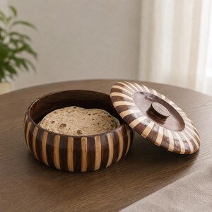 May include: A round wooden container with alternating dark brown and light tan vertical stripes. The container has a matching circular lid with a radial striped pattern and a central knob, containing a flat, round, beige food item inside.