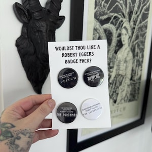 May include: A set of four circular pin-back badges on a white display card with the text "WOULDST THOU LIKE A ROBERT EGGERS BADGE PACK?". Three badges are black with white text, and one is white with black text, featuring film titles.
