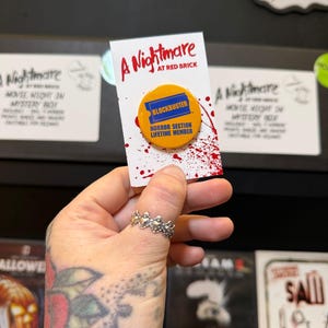 May include: A round yellow pinback button featuring a blue ticket graphic with the text BLOCKBUSTER and the words HORROR SECTION LIFETIME MEMBER. The button is attached to a white display card with the red text A Nightmare AT RED BRICK.
