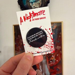 May include: A circular black pinback button featuring the white text I'D RATHER BE WATCHING GUILLERMO DEL TORO'S Frankenstein. The button is attached to a white display card with red blood splatter graphics and the text A Nightmare AT RED BRICK.