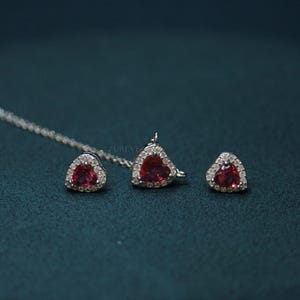Puede incluir: Un conjunto de joyería que consta de un colgante de collar plateado y dos pendientes a juego. Cada pieza presenta una piedra preciosa roja en forma de corazón rodeada por un halo de pequeñas piedras claras engastadas en un marco metálico.