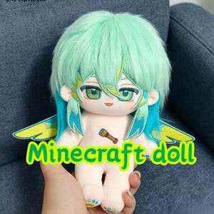 Puede incluir: Una muñeca de peluche con cabello verde menta, reflejos azul verdoso y ojos verdes. Presenta alas bordadas en amarillo y azul verdoso, un pequeño icono de telescopio marrón en el pecho y el texto "Minecraft doll" en letras verde brillante.