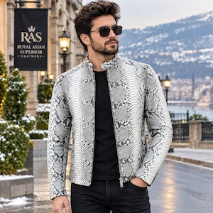 Può includere: Una giacca a maniche lunghe con motivo a pelle di serpente grigio e bianco, caratterizzata da un colletto alla coreana e una chiusura frontale con zip. La giacca è indossata sopra una maglia nera tinta unita e abbinata a pantaloni neri.