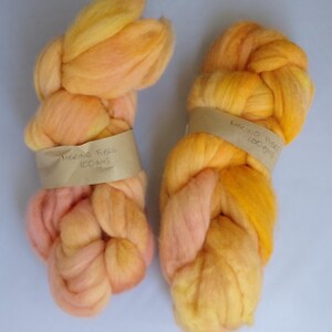 Puede incluir: Dos paquetes de lana merino suave sin hilar en tonos amarillo, naranja y melocotón. Cada paquete está sujeto con una banda de papel color canela con la etiqueta "MERINO FIBRE 100gms" escrita a mano en tinta negra, indicando el peso.