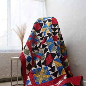 May include: A quilted blanket featuring a geometric pattern of stars and triangles in red, black, blue, yellow, and olive green. The fabric has a visible grid of stitched lines throughout, creating a textured surface across the entire textile piece.