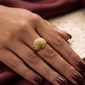 May include: A gold-toned ring featuring a fan-shaped design with raised, radiating lines that mimic the appearance of a seashell. The metal has a smooth, polished finish, and the ring is worn on the ring finger, showcasing its curved, semi-circular top.