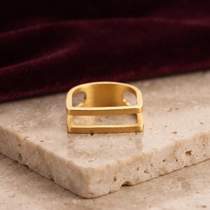 Può includere: Un anello color oro caratterizzato da un design geometrico con una barra superiore piatta e una barra inferiore parallela collegate da due supporti laterali. L'anello presenta una finitura opaca spazzolata e una struttura aperta dall'aspetto architettonico.