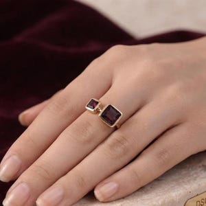 Puede incluir: Un anillo de oro con dos piedras preciosas de color rojo oscuro, de corte cuadrado y diferentes tamaños. La piedra más grande está engastada de forma prominente, mientras que la más pequeña se sitúa ligeramente desplazada en el aro.