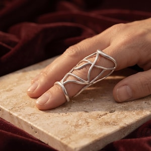 May include: A silver-toned finger splint featuring an open, cage-like lattice design that wraps around the finger. The structure consists of smooth, interconnected metal bands forming a series of irregular, elongated geometric shapes.