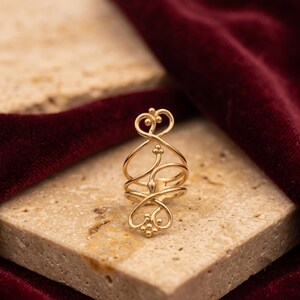 May include: A gold-toned ring featuring an elongated, openwork design with swirling wire patterns, delicate loops, and small spherical accents at the top and bottom terminals, creating a symmetrical, vertical structure that wraps around the finger.