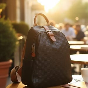 May include: A black backpack with a checkered pattern, featuring a tan leather top handle and a front strap closure with a metal buckle. The bag has a structured silhouette, a top carry handle, and adjustable shoulder straps in a matching tan color.