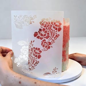 以下が含まれることがあります： 咲き誇る花々と葉のつるの複雑な切り抜きが施された、長方形の白いプラスチック製ステンシル。ステンシルは、同じ珊瑚色の浮き彫りの花柄が施された円筒形のケーキの表面に当てられています。