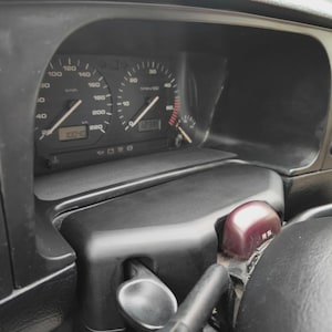 Puede incluir: Un panel de instrumentos de coche negro con velocímetro analógico en km/h, tacómetro y dos pantallas digitales. Un pequeño componente de plástico granate con el texto VW 104 es visible cerca de la columna de dirección.