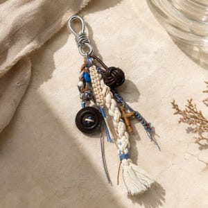 Puede incluir: Un llavero con un anillo de metal plateado unido a varios elementos decorativos, incluyendo un botón marrón oscuro, un cordón trenzado de color crema, una esfera anudada marrón oscuro, una pequeña cruz de madera y cuentas azules y blancas.
