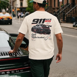Op de afbeelding: Een off-white t-shirt met een graphic op de achterkant van een grijze Porsche 911 GT3 RS. De tekst luidt 911 GT3 RS, Built for a Higher Standard, en bevat een kleine omtrek van een circuit. Het shirt wordt gedragen met een tweekleurige pet en donkere broek.
