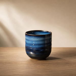 Puede incluir: Una taza de cerámica con un degradado de tonos azules, que transita desde un azul cielo claro en el borde hasta un azul marino profundo en la base. La superficie exterior presenta una serie de sutiles estrías horizontales texturizadas.