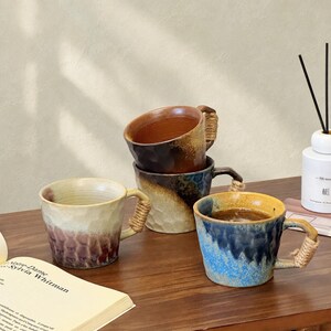 May include: Three ceramic mugs with textured, dimpled surfaces and wrapped rope handles. One mug is cream and purple, one is brown and tan, and one is blue and yellow. Each mug features a unique, reactive glaze finish and a sturdy, rounded handle.